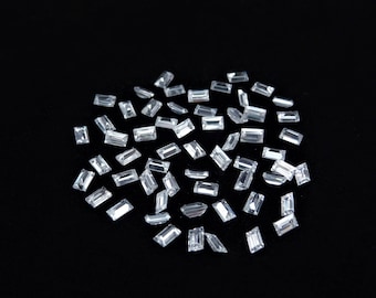 May include: A collection of numerous small, clear, rectangular baguette-cut gemstones scattered across a dark surface. Each individual stone features faceted edges that reflect light, creating a bright, transparent appearance throughout the group.