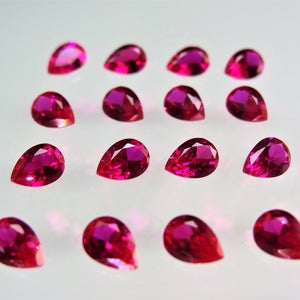 May include: A collection of 15 pear-shaped ruby gemstones, each with a deep red hue and a brilliant shine. The gemstones are arranged in a grid pattern on a white surface.