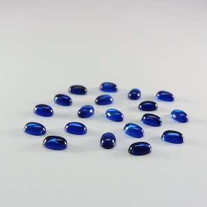 Puede incluir: Una colección de cabujones ovalados de vidrio azul dispuestos en círculo sobre una superficie blanca.