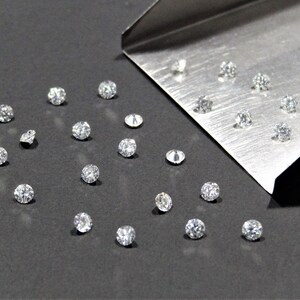 May include: A collection of loose, round, clear diamonds on a gray surface. Some diamonds are in a metal tray.