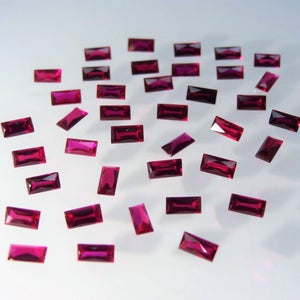 May include: A collection of 25 loose, rectangular, red gemstones. The gemstones are scattered on a white surface.