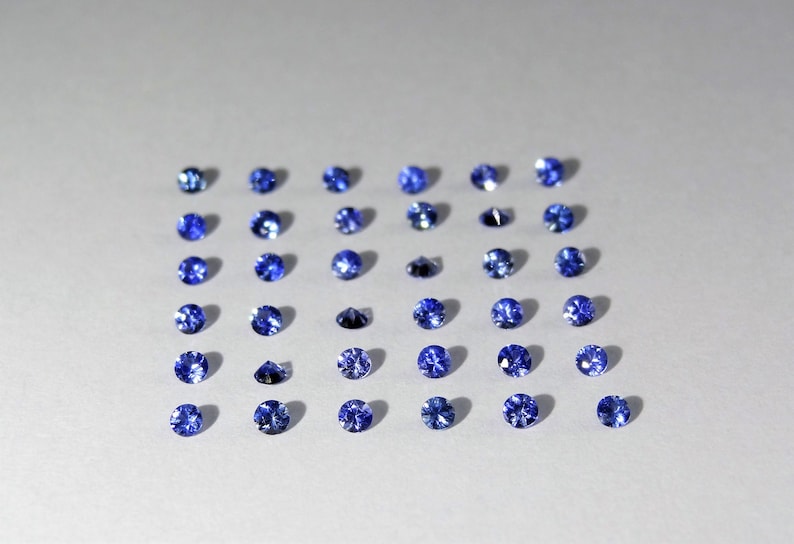May include: A collection of thirty small, round, faceted blue gemstones arranged in a grid of six rows and five columns. Each gemstone displays a brilliant cut with multiple reflective surfaces, showcasing a deep, vibrant blue color throughout.