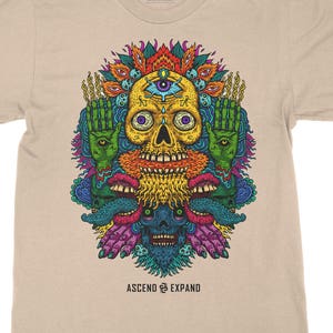 May include: A beige t-shirt with a colorful graphic print of a skull with a third eye, surrounded by hands and mouths. The text "ASCEND & EXPAND" is printed below the graphic.