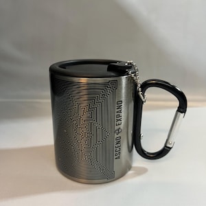 Puede incluir: Taza de viaje de acero inoxidable con tapa y asa negras. La taza presenta un diseño de laberinto geométrico y el texto "ASCEND EXPAND" impreso verticalmente. Una pequeña cadena está unida al asa.