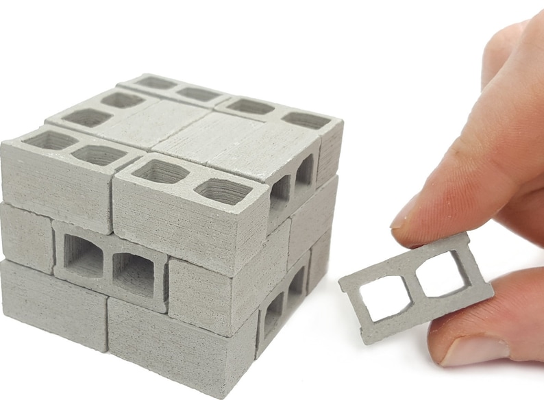 K&ouml;nnte beinhalten: Ein Satz Miniatur-Betonsteine, die zu einer kleinen Mauer gestapelt sind. Die Bl&ouml;cke sind grau und haben eine raue Textur. Ein einzelner Block wird in der Hand gehalten, wodurch die Details der L&ouml;cher sichtbar werden.