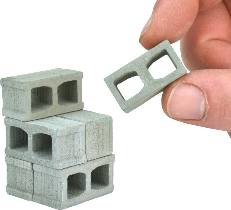 May include: A stack of four miniature concrete blocks, each with two rectangular holes, on a white background. A single block is held in a hand.