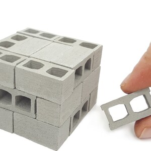 May include: A set of miniature concrete blocks, stacked to form a small wall. The blocks are gray and have a rough texture. A single block is held in a hand, showing the detail of the holes.