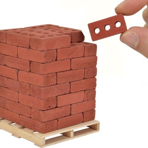 May include: A stack of miniature red bricks on a wooden pallet. The bricks are about the size of a fingertip and have a rough texture. A single brick is being held by a hand in the upper right corner of the image.