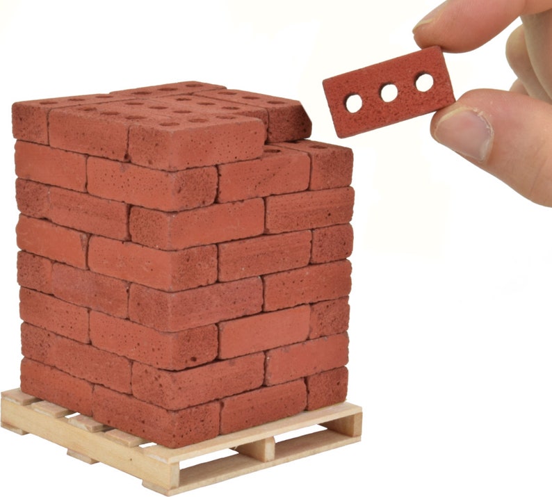 May include: A stack of miniature red bricks on a wooden pallet. The bricks are about the size of a fingertip and have a rough texture. A single brick is being held by a hand in the upper right corner of the image.