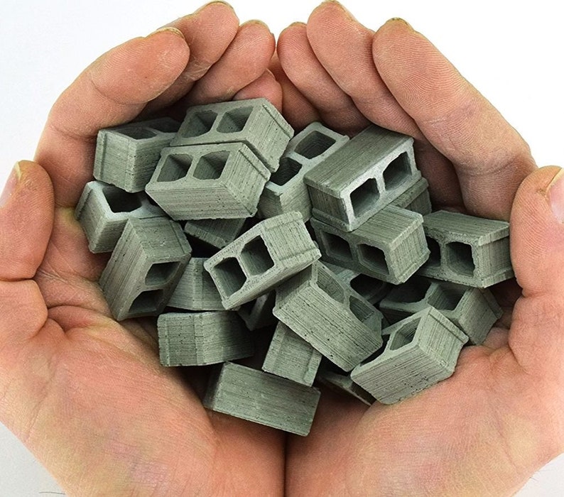 K&ouml;nnte beinhalten: Eine Handvoll Miniatur-Betonsteine, jeder mit einer hohlen Mitte, werden in den gefalteten H&auml;nden einer Person gezeigt. Die Bl&ouml;cke sind hellgrau und haben eine raue, strukturierte Oberfl&auml;che.