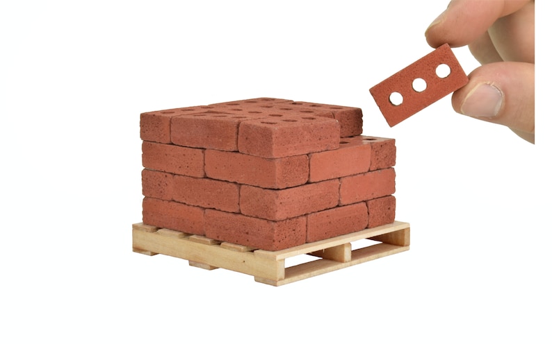 May include: A stack of red brick blocks on a wooden pallet. The bricks are arranged in a staggered pattern and have small holes in them. A single brick is being held by a hand, showing the holes in the brick.