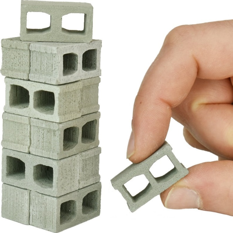 May include: A stack of eight miniature concrete blocks with two holes in each block. The blocks are gray and have a rough texture. A single block is being held by a hand.
