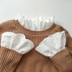 Puede incluir: Un suéter marrón acanalado con cuello y puños de encaje blanco. Los detalles de encaje presentan un borde festoneado, contrastando con la textura del suéter. Las mangas largas tienen puños de encaje que se extienden sobre las manos.