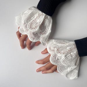 Può includere: Polsini in pizzo bianco con bordo smerlato e intricato design traforato. I polsini sono attaccati a una manica a coste nera. Un accessorio delicato.