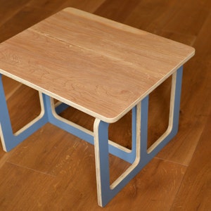 May include: A small, square wooden table with a light brown tabletop and blue legs. The table has a simple, modern design. The table measures approximately 46 cm long, 46 cm wide, and 30 cm high.