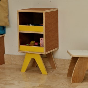 May include: A wooden storage cabinet with two yellow drawers and a matching yellow base. A small wooden chair with a white seat and back is next to the cabinet. The cabinet and chair are on a tiled floor.