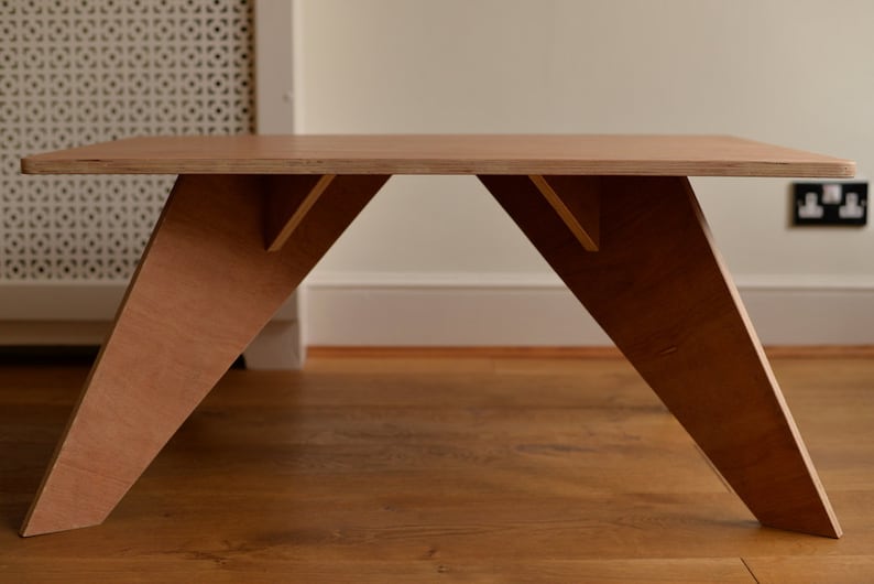 May include: A light brown wooden coffee table with a rectangular top and angled legs. The table has a minimalist design. The V-shaped legs provide stability. The table's dimensions are approximately 120 cm long, 60 cm wide, and 40 cm high.