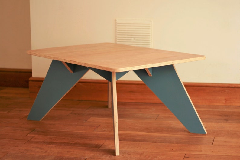 May include: A rectangular wooden coffee table with a light wood top and teal-coloured legs. The legs are angled, creating a modern design. The table is approximately 76 cm long and 51 cm wide.
