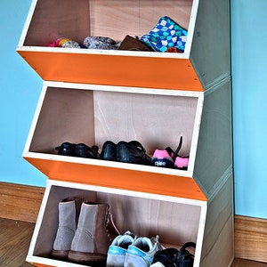 May include: A three-tiered wooden storage unit with an orange front and light wood interior. The unit is filled with shoes, boots, and other items. The unit is angled and has a modern design. The background is a light blue wall.
