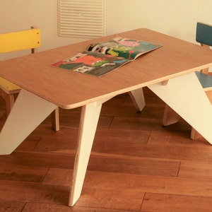 May include: A children's table and two chairs. The table has a light wood top and white angled legs. One chair has a yellow back and seat, the other has a blue back and seat. A book is open on the table. Table dimensions are approximately 75cm long.