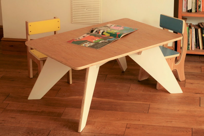 May include: A children's table and two chairs. The table has a light wood top and white angled legs. One chair has a yellow back and seat, the other has a blue back and seat. A book is open on the table. Table dimensions are approximately 75cm long.
