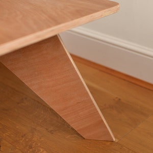 May include: Close-up of a light brown wooden table with an angled leg design. The table's surface and leg are made of the same wood, showing the natural grain. The table is on a wooden floor.