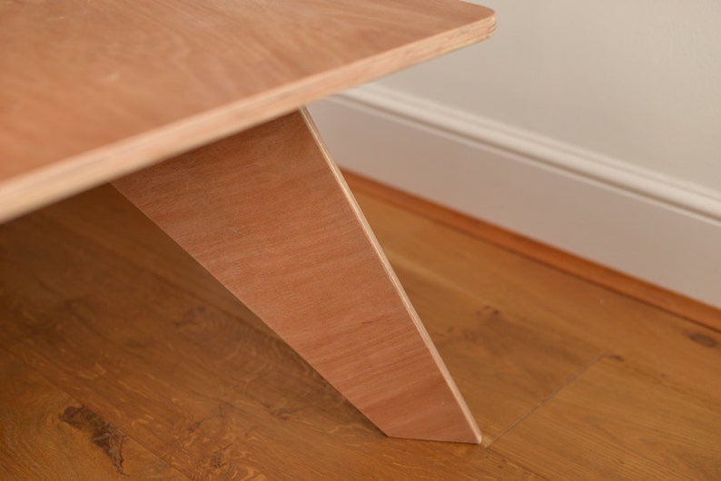 May include: Close-up of a light brown wooden table with an angled leg design. The table's surface and leg are made of the same wood, showing the natural grain. The table is on a wooden floor.