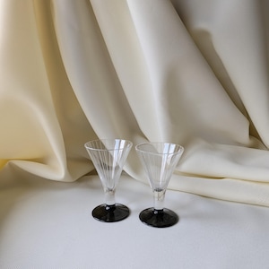 May include: Two clear, stemmed shot glasses with a conical design and black bases. The glasses have vertical lines etched into the glass, creating a textured effect. They are set against a cream-colored fabric background.