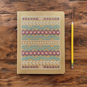 May include: A brown notebook with a colorful geometric pattern stitched on the cover. The notebook is lying on a wooden surface next to a yellow pencil.