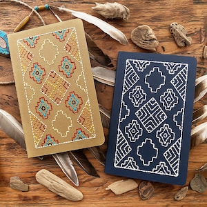 May include: Two notebooks with geometric patterns stitched on the covers. One notebook has a tan cover with colorful stitching, and the other has a navy blue cover with white stitching.