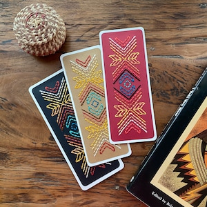 May include: Three handmade bookmarks with geometric patterns. The bookmarks are made with colorful embroidery thread on a variety of fabric backgrounds. The bookmarks are red, brown, and black.
