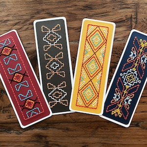May include: Four colorful bookmarks with geometric patterns embroidered on them. The bookmarks are on a wooden surface.