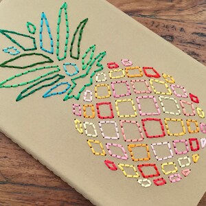May include: A brown notebook with a colorful embroidered pineapple design. The pineapple is outlined in black thread and filled with squares of different colors, including red, orange, yellow, green, blue, and pink.