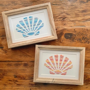 May include: Two framed cross-stitch seashell art pieces. One features blue and white hues, the other red, orange, and yellow. Both are in light wood frames, set against a wooden background.