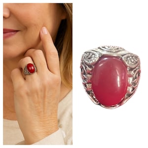 May include: A silver-toned ring with an oval, red gemstone. The ring has an ornate, engraved design around the stone. The image also shows a person wearing the ring on their finger.