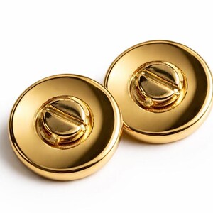 May include: Two gold-colored, round objects with a screw-like design in the center. The objects have a shiny, reflective surface, and are set against a white background. The design is a close-up shot.