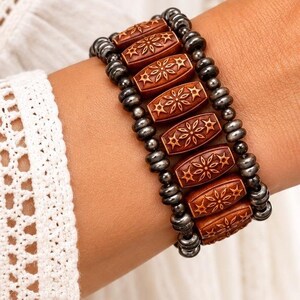 May include: A multi-strand bracelet featuring two rows of small, metallic dark gray beads flanking a central row of six rectangular, polished brown wooden beads, each engraved with a symmetrical floral star pattern and rounded edges.