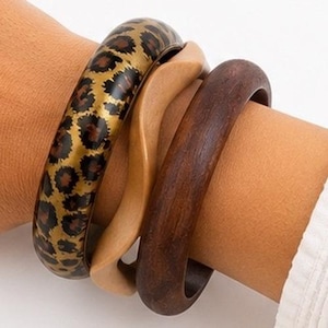May include: A set of three bangles consisting of one gold-toned bangle with a black and brown leopard print pattern, one light tan wavy-edged bangle, and one dark brown smooth bangle, all worn together on a wrist.