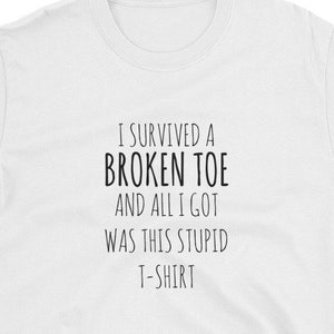 Op de afbeelding: Wit t-shirt met de tekst "I SURVIVED A BROKEN TOE AND ALL I GOT WAS THIS STUPID T-SHIRT" in het zwart. Het shirt heeft korte mouwen en een ronde hals. De tekst staat in het midden van de voorkant.