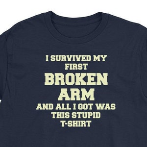 Pode incluir: Camiseta azul marinho com o texto "I SURVIVED MY FIRST BROKEN ARM AND ALL I GOT WAS THIS STUPID T-SHIRT" em negrito, fonte creme. A camiseta é de manga curta, adequada para uso casual.