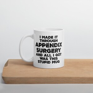 May include: White ceramic mug with the text "I MADE IT THROUGH APPENDIX SURGERY AND ALL I GOT WAS THIS STUPID MUG" in black. The mug is on a wooden surface.