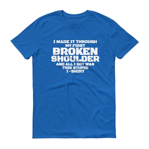 Op de afbeelding: Een koningsblauw t-shirt met witte tekst. De tekst luidt: "IK HEB MIJN EERSTE GEBROKEN SCHOUDER DOORSTONDEN EN ALLES WAT IK KREEG WAS DIT STOMME T-SHIRT."
