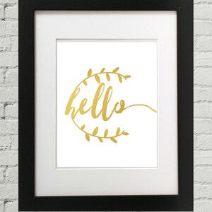 Puede incluir: Una imagen enmarcada en negro con un passe-partout blanco y una impresión en lámina dorada de la palabra "hello" rodeada de una corona de hojas en lámina dorada.