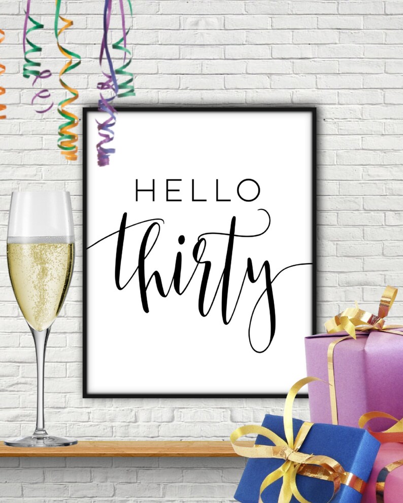 Puede incluir: Un cartel imprimible en blanco y negro con el texto "Hello thirty" en una fuente de letra cursiva decorativa. El cartel est&aacute; enmarcado en un marco negro y est&aacute; sentado en un estante de madera con una copa de champ&aacute;n y regalos envueltos con cinta dorada.