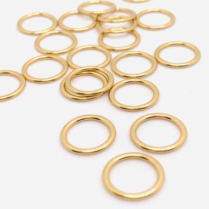 May include: A group of gold jump rings scattered on a white surface. The rings are all the same size and are made of a shiny metal.