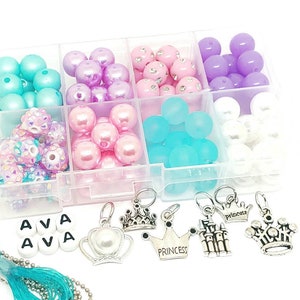 Puede incluir: Un organizador de plástico transparente lleno de cuentas de colores en tonos rosa, morado, azul y blanco. Incluye dijes de corona plateada, cuentas de letras que deletrean "AVA" e hilo turquesa para hacer joyas.