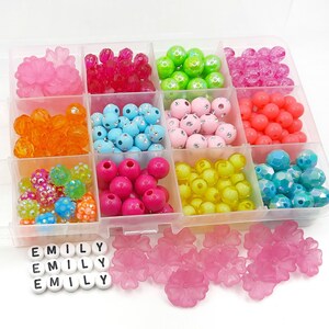 May include: A clear plastic organizer filled with various colorful beads and embellishments. The beads are in shades of pink, orange, green, blue, and yellow. Also included are pink flower-shaped beads and letter beads spelling "EMILY".