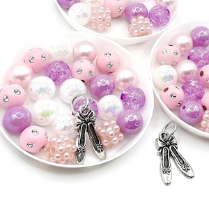 May include: A collection of pink and purple beads with silver ballerina shoe charms. The beads are arranged on white dishes.