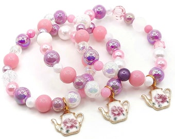Op de afbeelding: Drie roze en paarse kralenarmbanden met theepot bedels. De bedels zijn goudkleurig met een wit bloemenontwerp.