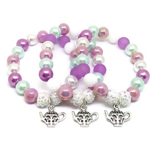 May include: Three beaded bracelets with iridescent pastel purple, white, and green beads. Each bracelet has a silver charm shaped like a teapot.